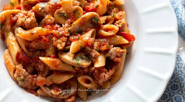 Piatto di pasta alla boscaiola con penne, funghi, salsiccia e pomodoro, guarnito con prezzemolo.