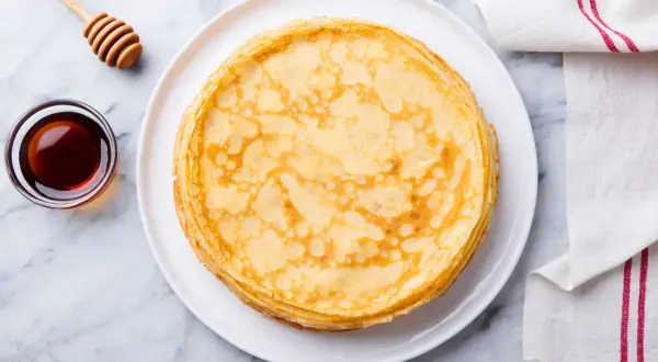 Pila di crepes senza uova dorate su un piatto bianco, con una ciotola di sciroppo d'acero e un dosatore di miele accanto.