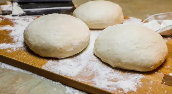 Tre panetti soffici di pizza a lunga lievitazione, pronti per essere stesi su un tagliere di legno infarinato.