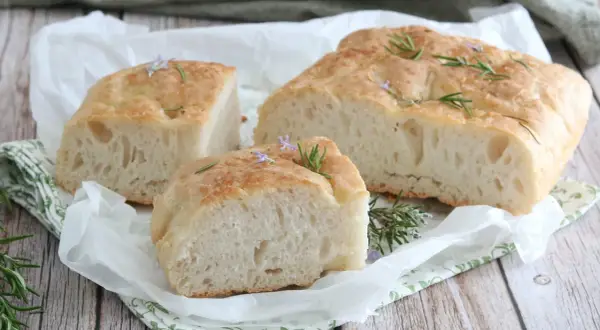 Focaccia senza glutine soffice e dorata, guarnita con rosmarino fresco e piccoli fiori viola, su carta da forno.