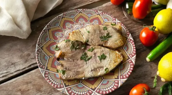Due fette di pesce spada in friggitrice ad aria, guarnite con prezzemolo, su un piatto decorato. Accanto, pomodori, limoni e zucchine.