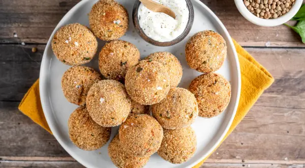 Piatto di polpette di lenticchie dorate e croccanti, servite con una ciotolina di salsa cremosa e un cucchiaino di legno.