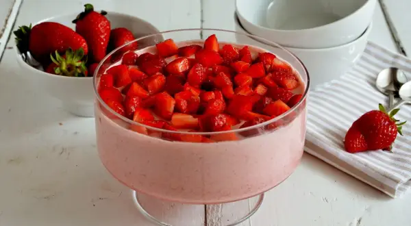Delizioso mousse di fragole in una coppa di vetro, guarnito con fragole fresche a pezzi. Accanto, una ciotola di fragole e ciotole bianche.