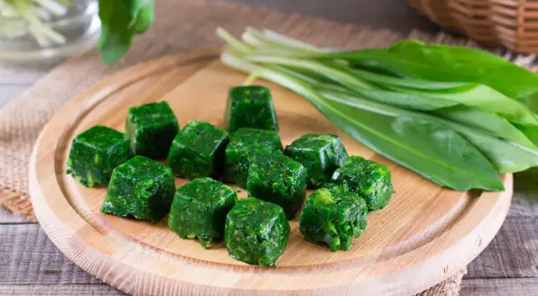 Cubetti di spinaci surgelati in padella, pronti per essere cucinati. Accanto, un mazzo di spinaci freschi.