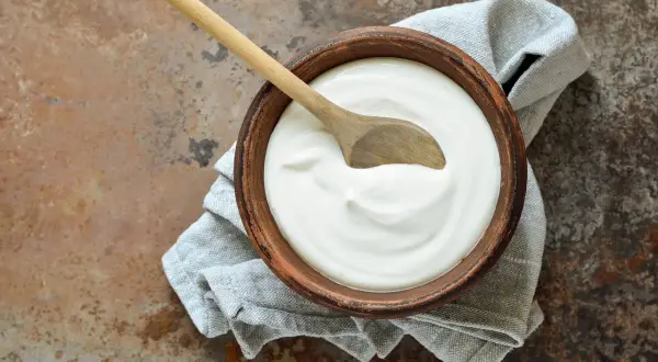 Ciotola di terracotta con cremoso yogurt fatto in casa, un cucchiaio di legno appoggiato sopra, su un tovagliolo grigio.