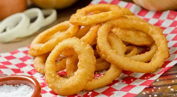 Anelli di cipolla dorati e croccanti, preparati con una perfetta pastella per fritti, serviti su carta a scacchi rossi e bianchi, con sale grosso a lato.