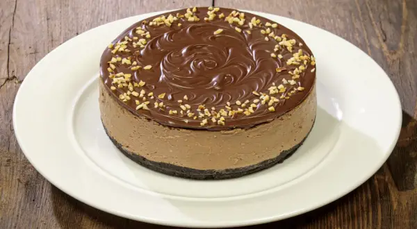 Cheesecake alla Nutella con glassa al cioccolato e nocciole tritate, su un piatto bianco.
