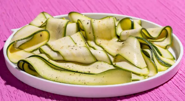 Fettine sottili di zucchine marinate crude, condite con pepe nero, disposte a spirale in un piatto bianco su un tavolo rosa acceso.