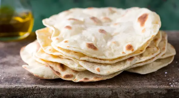 Pila di pane senza lievito, cotto su una tavola di legno grezzo, con un tocco di sale e una bottiglia d'olio sullo sfondo.