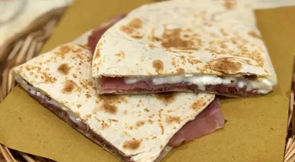 Piadina fatta in casa con prosciutto e formaggio filante, servita su carta gialla in un cestino di vimini.