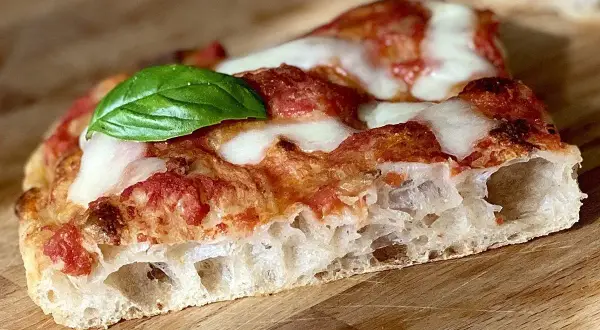 Fetta di pizza in teglia con pomodoro, mozzarella filante e una foglia di basilico fresco. La crosta è alta e alveolata, segno di una lievitazione perfetta.