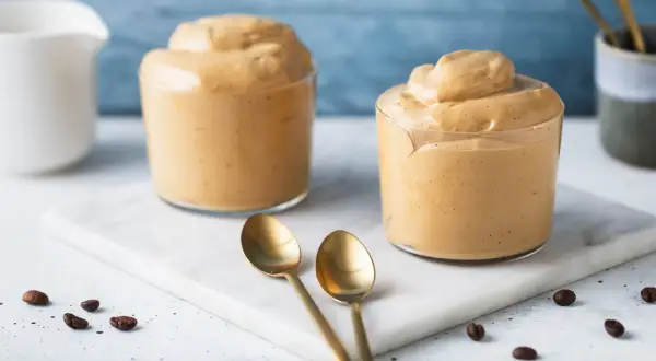 Due bicchieri di cremosa crema di caffè senza panna, perfetti per una pausa golosa. Chicchi di caffè e cucchiaini dorati completano la scena.