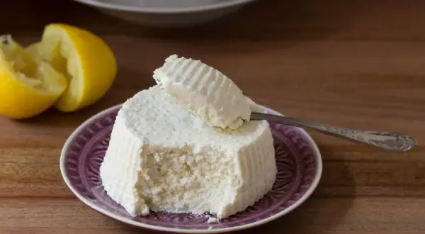 Ricotta fatta in casa su un piatto viola, con limone a fianco. Un cucchiaino preleva una porzione cremosa.