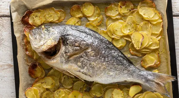 Orata al forno dorata e succulenta, adagiata su un letto di patate affettate e cotte a puntino, profumate di erbe aromatiche.