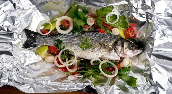 Branzino al cartoccio pronto per la cottura, con pomodorini, cipolla e prezzemolo su foglio d'alluminio.