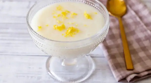 Deliziosa crema di riso in una coppa di vetro, guarnita con scorza di limone. Accanto, un cucchiaio dorato e un tovagliolo a quadretti.
