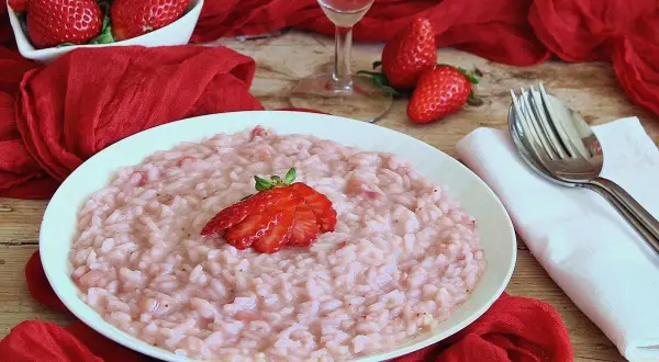 Un cremoso risotto alle fragole, guarnito con una fragola fresca, su un piatto bianco, circondato da fragole rosse e un drappo di seta.