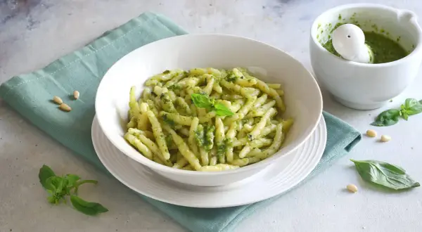 Un piatto invitante di pasta al pesto, condita con basilico fresco e pinoli, accanto a un mortaio con altro pesto.