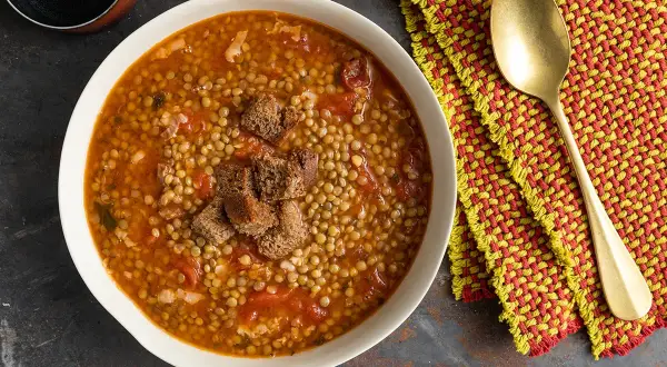 Una ciotola di minestra di lenticchie fumante, guarnita con crostini di pane, accanto a un tovagliolo colorato e un cucchiaio dorato.