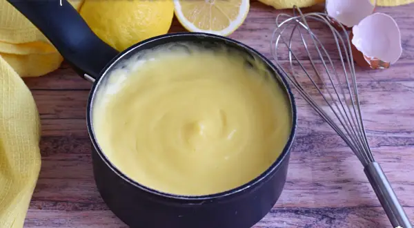 Crema pasticcera liscia e vellutata, preparata con amido di mais, pronta per farcire dolci. Accanto, limoni e frusta.