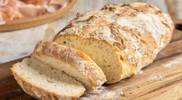 Pane fatto in casa con farina 00, tagliato a fette su un tagliere di legno, con affettati in lontananza.