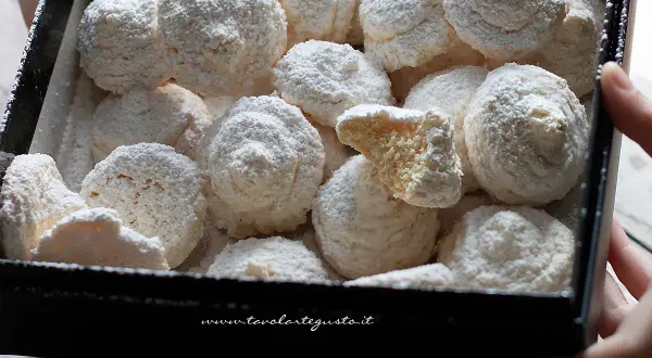 Scatola piena di soffici biscotti al cocco, spolverati di zucchero a velo. Uno è spezzato, rivelando la sua consistenza friabile.