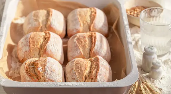 Sei panini dorati, pronti per essere cotti nel pane in friggitrice ad aria. La farina spolverata e le spighe di grano completano la scena.