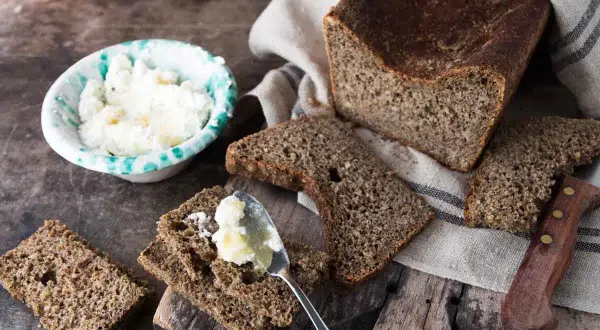 Fetta di pane di segale con burro spalmato, accanto a una ciotola di formaggio fresco e un coltello.