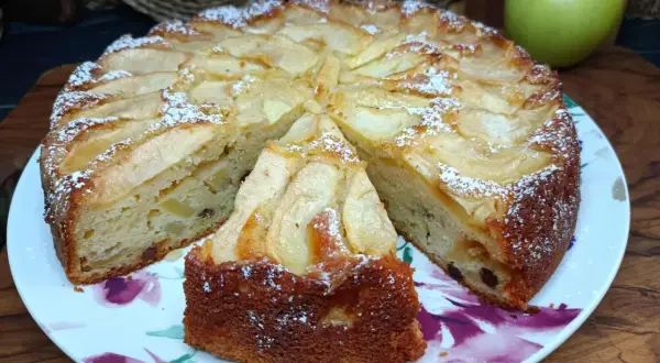Una fetta di torta di mele, cosparsa di zucchero a velo, su un piatto fiorato. Sullo sfondo, una mela verde.