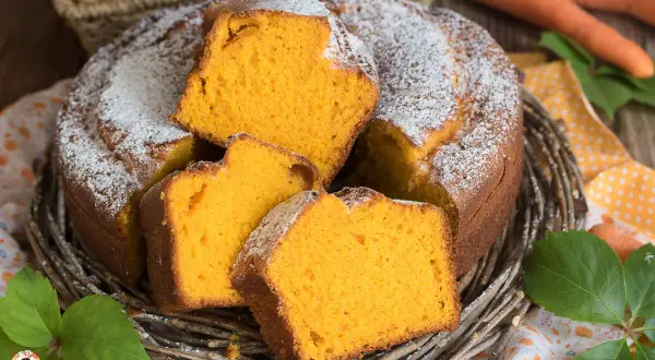 Una soffice torta di carote, cosparsa di zucchero a velo, servita in un cestino di vimini. Le fette mostrano l'interno arancione e invitante.