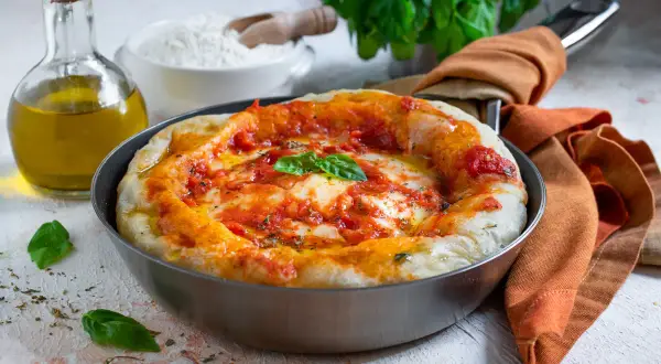 Pizza in padella appena sfornata, con pomodoro, mozzarella filante e basilico fresco. Accanto, olio d'oliva e farina, ingredienti per un'ottima pizza fatta in casa.