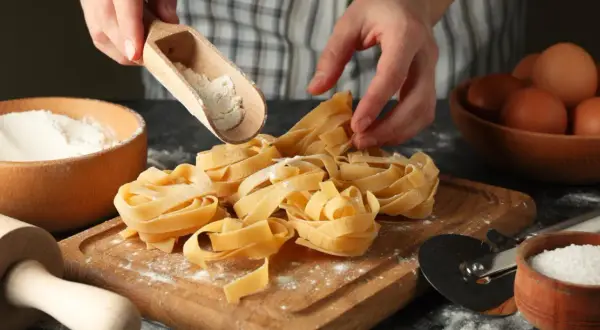 Mani che spolverano farina su nidi di pasta fatta in casa. Ingredienti freschi, uova e sale, pronti per un pasto delizioso.