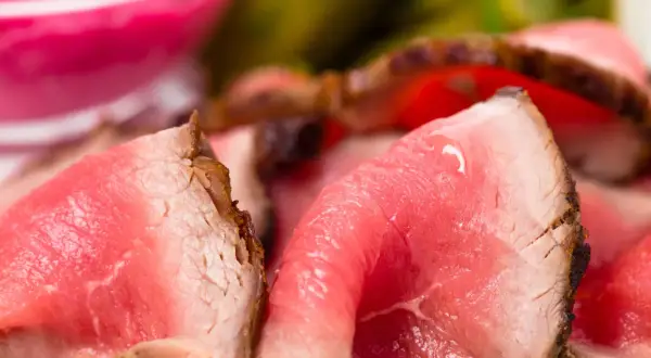 Fette sottili di roast beef ricetta classica, succulente e rosate, con un bordo croccante. Sullo sfondo, un contorno rosa e verde sfocato.