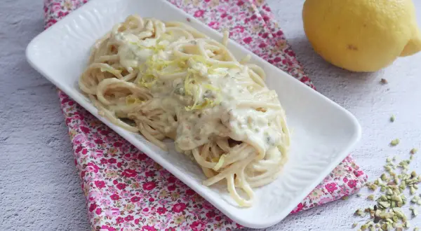 Un piatto di spaghetti al limone cremosi, guarniti con scorza di limone e pistacchi tritati, accanto a un limone intero.