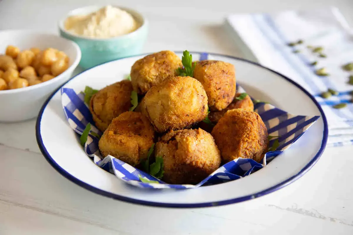 Un piatto di polpette di ceci dorate e croccanti, guarnite con prezzemolo fresco. Sullo sfondo, ciotole con ceci e una crema chiara.