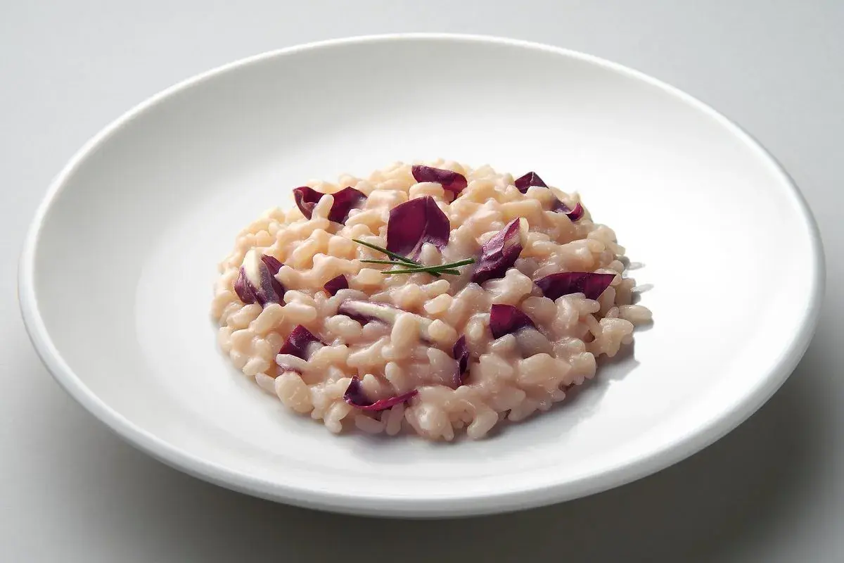 Un cremoso risotto al radicchio, guarnito con foglie viola e un rametto di erba cipollina, servito su un piatto bianco.