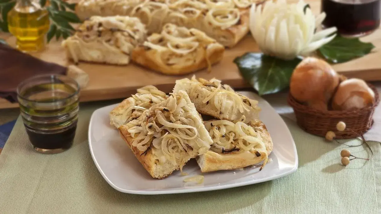Focaccia con cipolle dorate, fragrante e invitante, servita su un piatto bianco. Sullo sfondo, ingredienti freschi e una bottiglia d'olio.