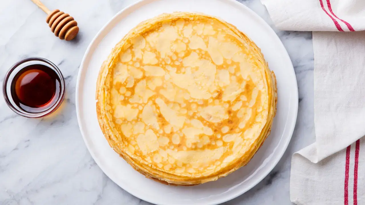 Pila di crepes senza uova dorate su un piatto bianco, con una ciotola di sciroppo d'acero e un dosatore di miele accanto.