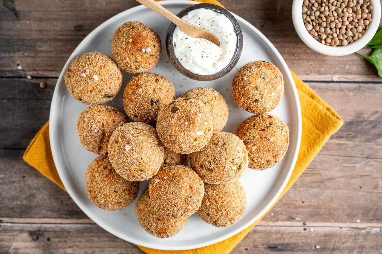 Piatto di polpette di lenticchie dorate e croccanti, servite con una ciotolina di salsa cremosa e un cucchiaino di legno.