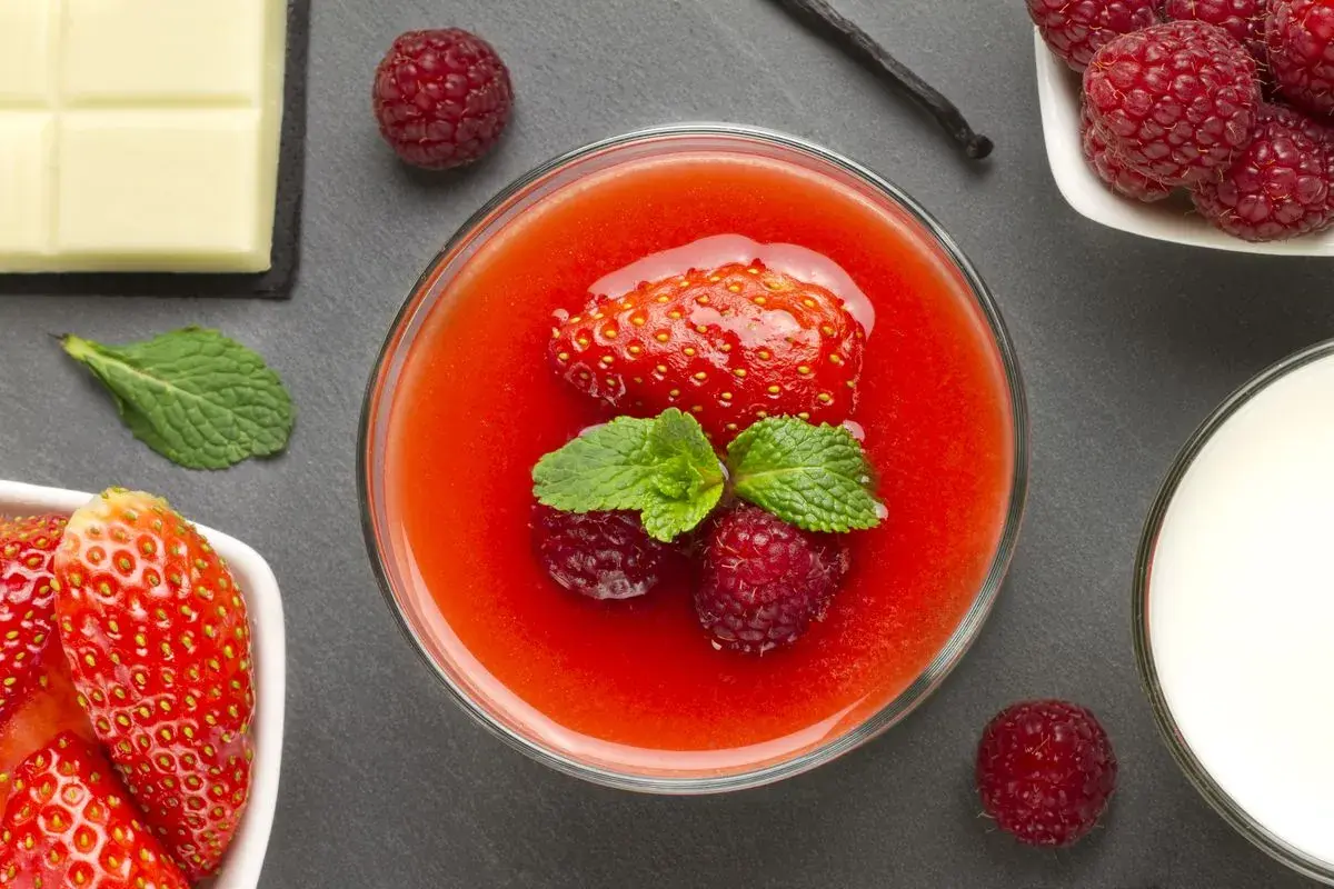 Un bicchiere di coulis di fragole con fragola, lamponi e menta. Accanto, cioccolato bianco, lamponi freschi e un bicchiere di latte.