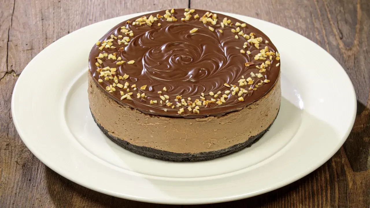 Cheesecake alla Nutella con glassa al cioccolato e nocciole tritate, su un piatto bianco.