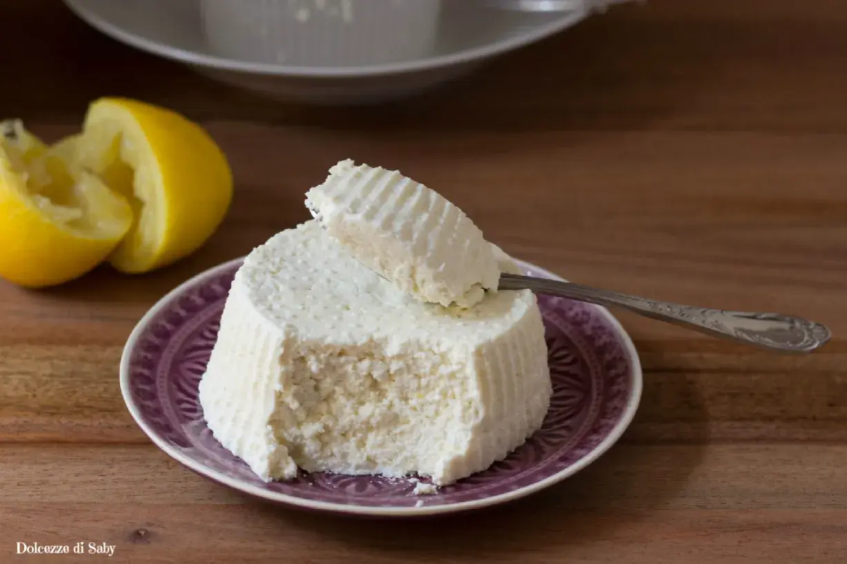 Ricotta fatta in casa su un piatto viola, con limone a fianco. Un cucchiaino preleva una porzione cremosa.