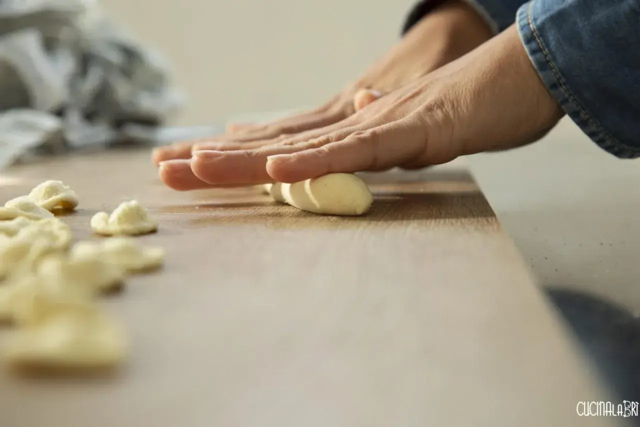 Mani che modellano impasto per creare orecchiette fatte in casa su un tagliere di legno.