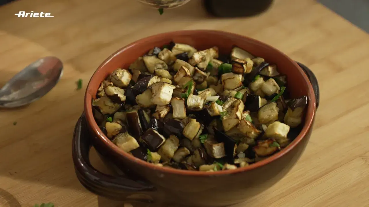 Melanzane in friggitrice ad aria, tagliate a cubetti e condite con erbe aromatiche, servite in una ciotola di terracotta.