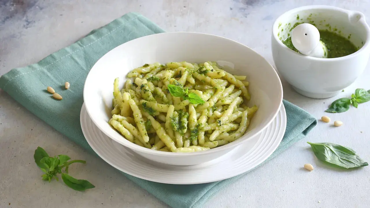 Un piatto invitante di pasta al pesto, condita con basilico fresco e pinoli, accanto a un mortaio con altro pesto.