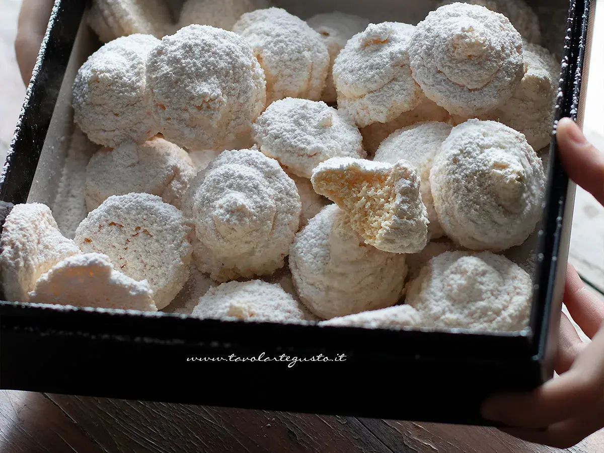 Scatola piena di soffici biscotti al cocco, spolverati di zucchero a velo. Uno è spezzato, rivelando la sua consistenza friabile.
