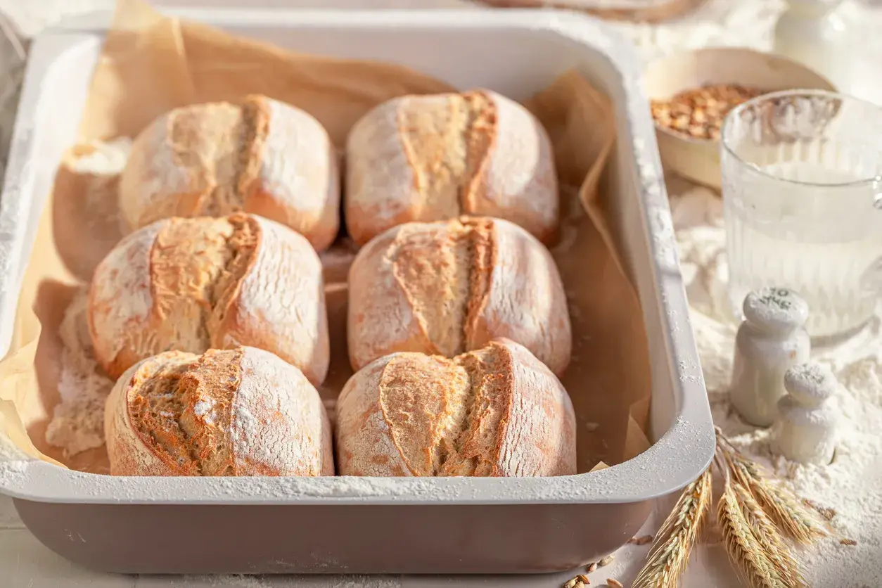 Sei panini dorati, pronti per essere cotti nel pane in friggitrice ad aria. La farina spolverata e le spighe di grano completano la scena.