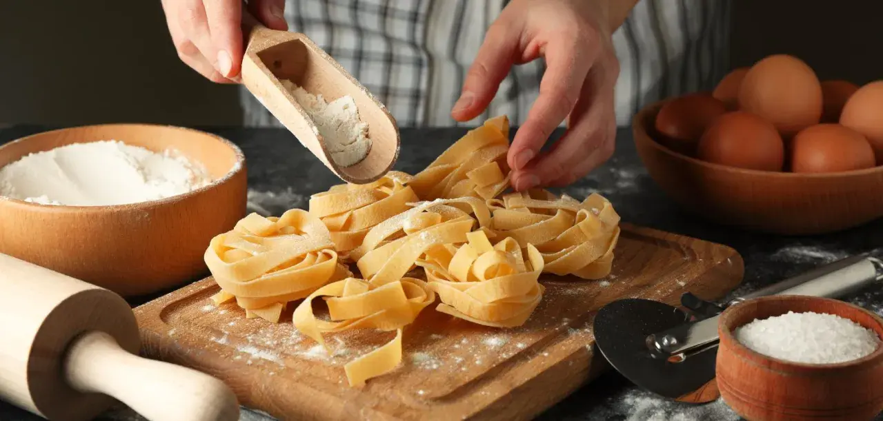 Mani che spolverano farina su nidi di pasta fatta in casa. Ingredienti freschi, uova e sale, pronti per un pasto delizioso.