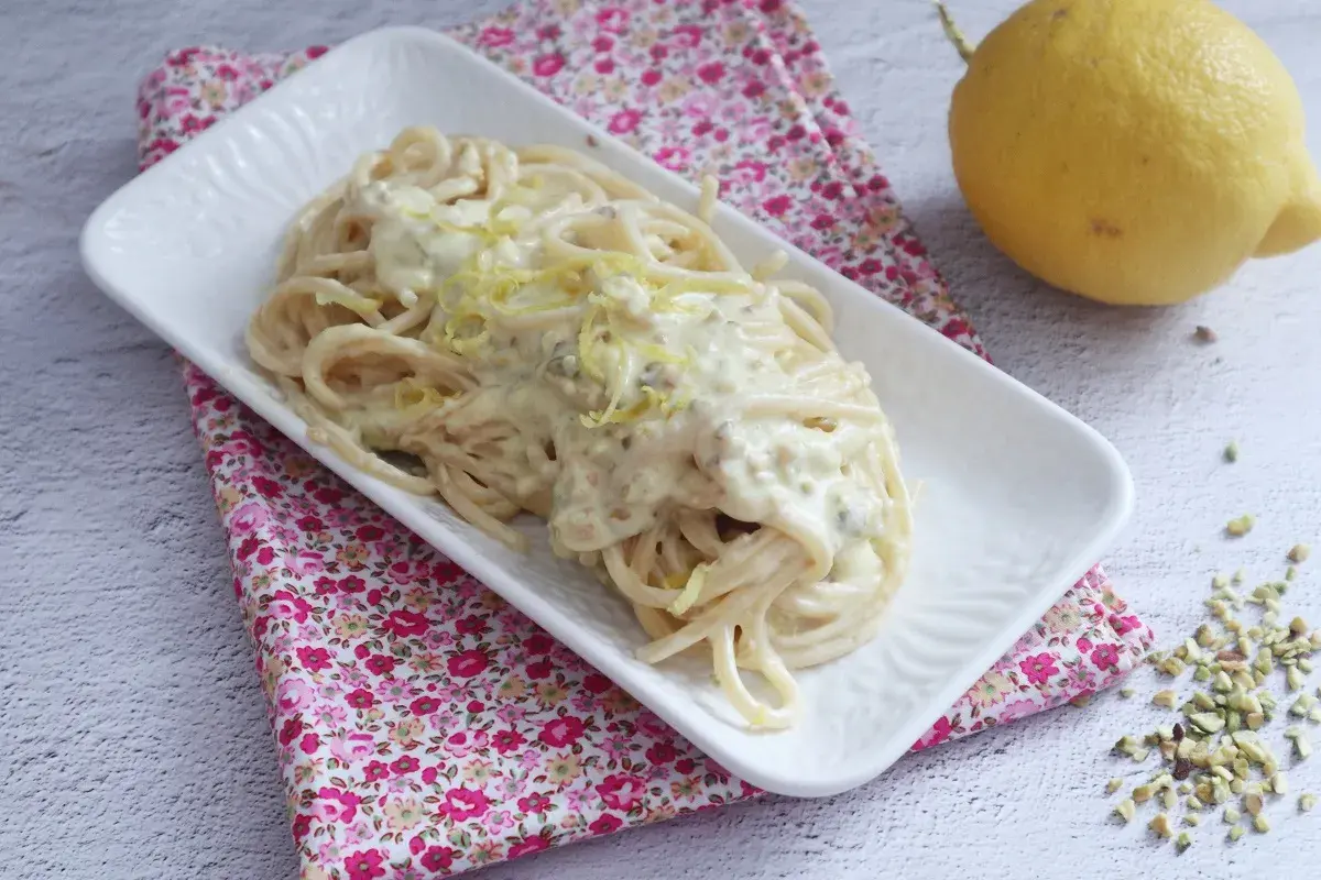 Un piatto di spaghetti al limone cremosi, guarniti con scorza di limone e pistacchi tritati, accanto a un limone intero.