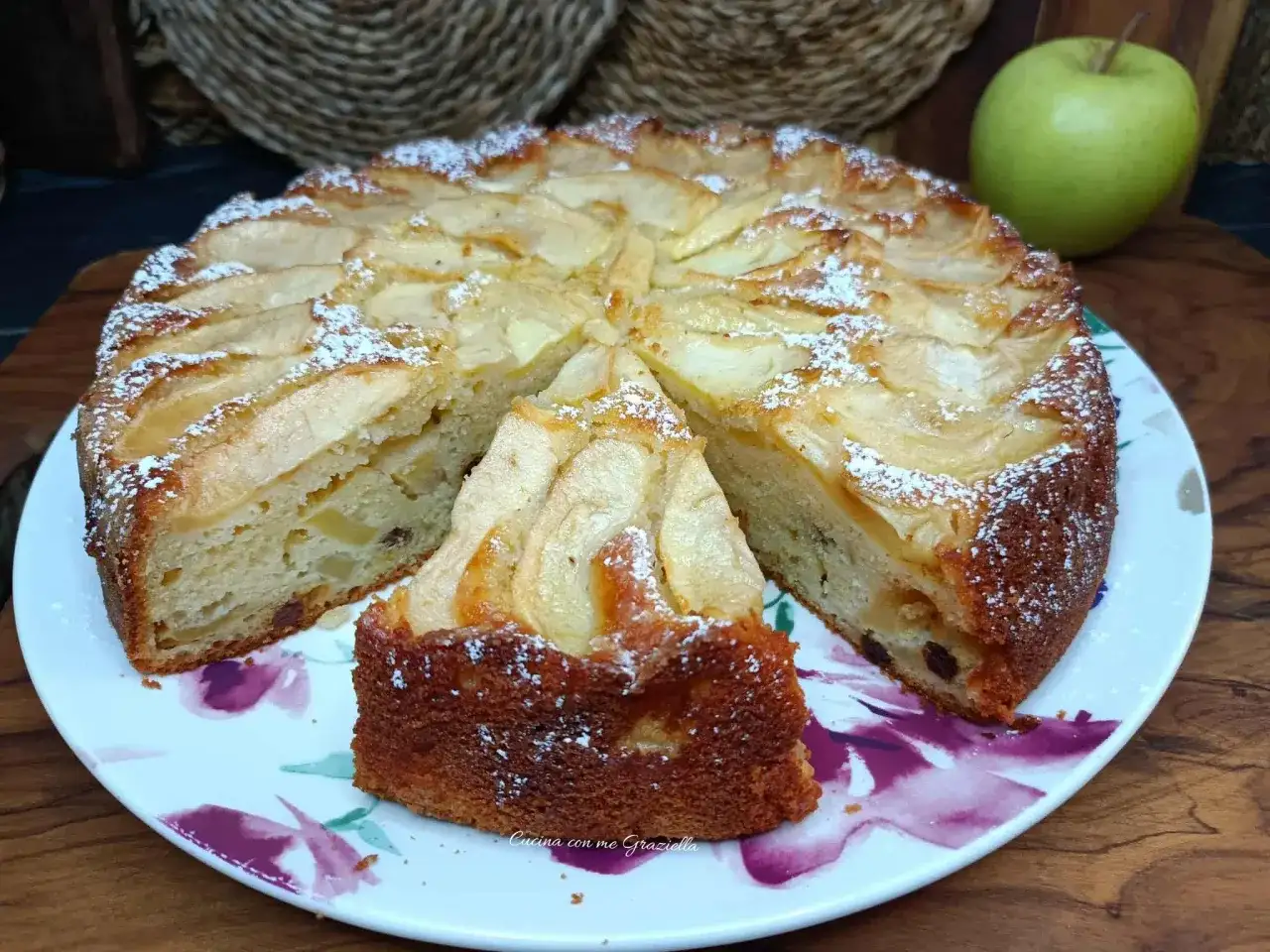 Una fetta di torta di mele, cosparsa di zucchero a velo, su un piatto fiorato. Sullo sfondo, una mela verde.